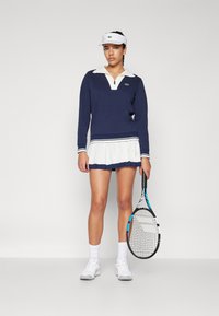 Marinblå tröja med vit krage och kant, kombinerad med en vit veckad kjol. Modellen håller ett tennisracket och har på sig vita skor och en visir.