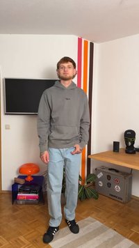Siva kapuca s prednjim dzepom, svijetloplave traperice i crne tenisice. Prostorija ima šareni zid, televizor i namještaj.