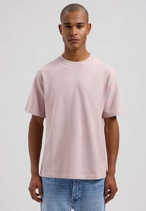 Mand står og vender fremad iført en lyserød T-shirt med korte ærmer og blå jeans foran en ensfarvet baggrund.