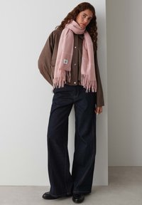 Light pink scarf with fringe, worn over a brown jacket with press studs, paired with dark wide-leg jeans and black shoes.