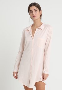 Abito camicia rosa chiaro a maniche lunghe con chiusura a bottoni, colletto e una tasca sul petto. Tessuto morbido con vestibilità ampia e orlo curvo.