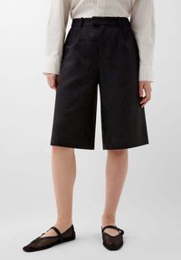 Shorts noirs arrivant jusqu'au genou en tissu texturé, dotés d'une taille haute, de passants de ceinture et d'une coupe droite. Associés à des ballerines noires translucides.