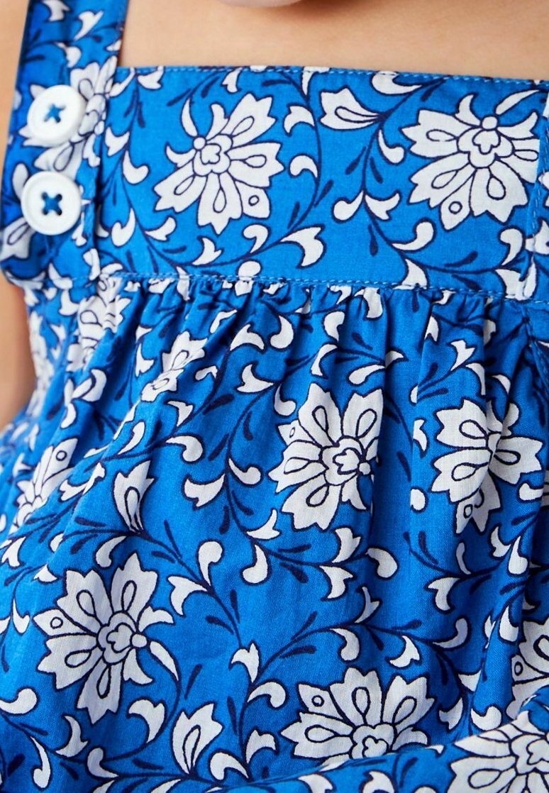 Sininen kukkamekko valkoisilla kukkakuvioilla, jossa on kerroshame, leveät hihnat ja kaksi valkoista nappia vartalolla.