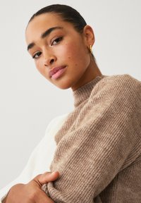 Maglione marrone a maglia con colletto alto e texture rigata, dotato di maniche lunghe. Una mano tiene una parte del maglione; sfondo chiaro.