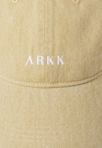 Gorra caqui hecha de tela texturizada con visera curvada y logo bordado en blanco "ARKK" en la parte frontal. Detalle de costura visible en la corona.