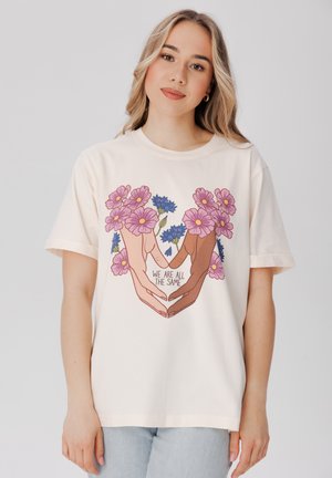 Camiseta de algodón color crema con un gráfico de dos manos formando un corazón lleno de flores rosas y azules, con el texto "TODOS SOMOS IGUALES."