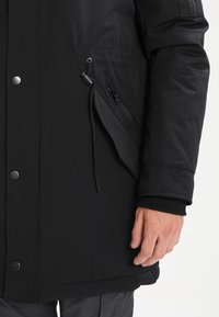 Svart isolerad jacka med slät tyg, tryckknapp- och blixtlåsstängning samt en sidoficka med blixtlås. Manschetten är ribbad för bättre passform.