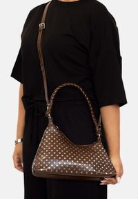 Bolso de mano de cuero marrón con textura suave, que presenta un asa trenzada y patrones de puntos plateados, llevado al hombro con una correa ajustable.