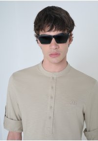 Camicetta corta color beige con placca a bottoni e maniche arrotolate, caratterizzata da un logo ricamato discreto. Occhiali da sole scuri con forma rettangolare.