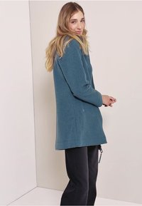 Veste en polaire teal, présentant un design mi-long avec des poches latérales et une coupe décontractée. Portée sur un pantalon noir.