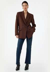 Blazer doppio petto marrone in tessuto strutturato con due tasche frontali, abbinato a jeans svasati blu scuro e stivaletti neri.