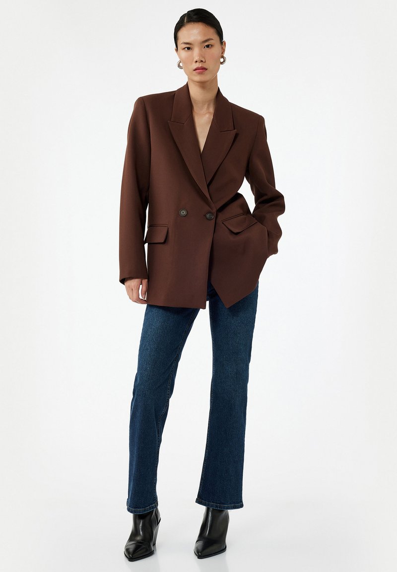 Blazer doppio petto marrone in tessuto strutturato con due tasche frontali, abbinato a jeans svasati blu scuro e stivaletti neri.