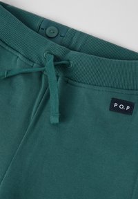 Teal-hihna housut, joissa on ribattu vyötärö, ommeltu napin sulkeminen vyötärön sisäpuolella ja pieni musta etiketti, jossa on valkoiset "P.O.P"-kirjaimet.