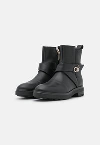 Botas de tobillo de cuero negro con suela gruesa, cierre de cremallera lateral y una amplia correa negra con un detalle de logo metálico.