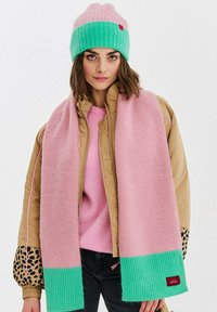 Pletená čiapka v ružovej a zelenej farbe, doplnená ružovým šálom so zeleným rebrovaným okrajom. Modelka má na sebe hnedú bundu s leopardím vzorom.