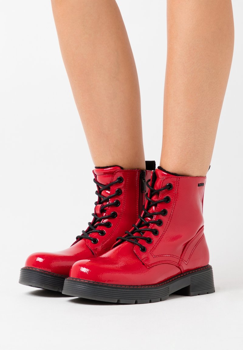 Bottes de cheville rouges brillantes à lacets avec semelles noires sur des jambes nues, dotées de fermetures éclair latérales et de talons épais à semelles crantées.