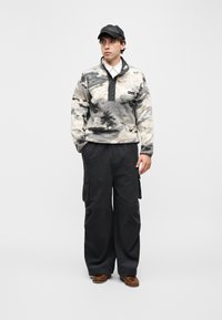 Νεαρός άνδρας που στέκεται, φορώντας fleece πουλόβερ με μοτίβο, φαρδιά μαύρη cargo παντελόνια, καφέ παπούτσια, λευκό πουκάμισο και μαύρο δερμάτινο καπέλο baseball.