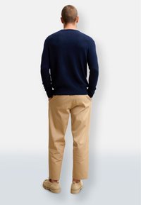 Marinblå stickad tröja med rundad halsringning, beiga bomullsbyxor och ljusa skor, sedd bakifrån mot en neutral bakgrund.