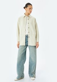 Giacca color crema con colletto, chiusura con bottoni e due tasche frontali, abbinata a jeans a gamba larga di un leggero blu chiaro. Design semplice.