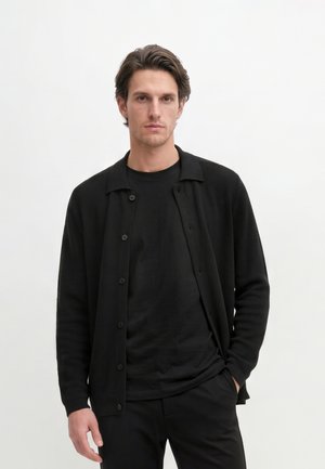 Jonge man met donker haar die een zwarte gebuttonde cardigan draagt over een zwart overhemd, staand tegen een effen witte achtergrond.