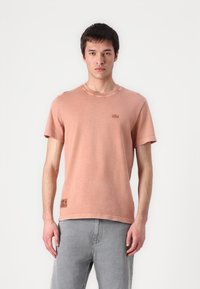 Tricou roz cu mânecă scurtă, confecționat din bumbac, cu decolteu rotund și un mic logo brodat pe piept. Puratat cu blugi gri.