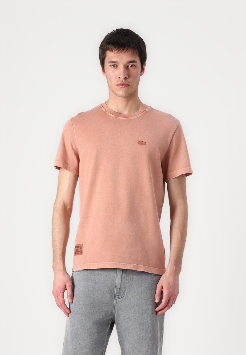 Rožinė trumpa rankovė t-shirt, pagaminta iš medvilnės, su apvalia iškirpte ir mažu logotipu, išsiuvinėtus ant krūtinės. Dėvima su pilkais džinsais.