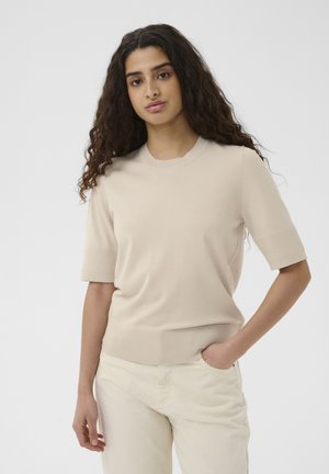 Junge Frau mit langen, lockigen Haaren, die einen beigen Kurzarmpullover und hellfarbene Hosen trägt und mit einer Hand in der Tasche steht.