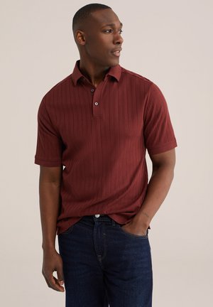 Muž nosiaci vínovú žerzejovú polokošeľu s krátkym rukávom a tmavomodré rifle, pozerá sa doprava s jednou rukou v vrecku.