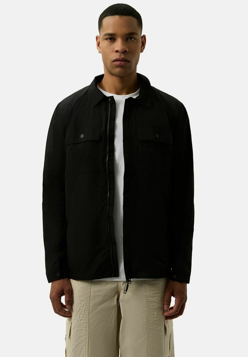 Zwarte lichte jas met een knoopsluiting aan de voorkant, twee borstzakken en een zachte textuur. Draagt over een wit T-shirt en beige broek.