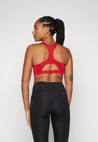 Sostenedor deportivo rojo con un diseño de espalda nadadora y detalle recortado. La modelo lleva leggings negros de cintura alta, mostrando texturas suaves.