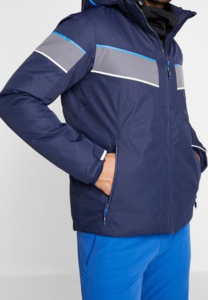 Homme portant une veste d'hiver bleu marine avec des rayures grises et bleues, fermée par une fermeture éclair, les mains dans les poches latérales, assortie à un pantalon bleu vif.