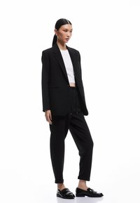 Sort skræddersyet blazer over en hvid cropped top, parret med løse sorte bukser og blanke sorte loafers. Glat stof med minimal detaljer.