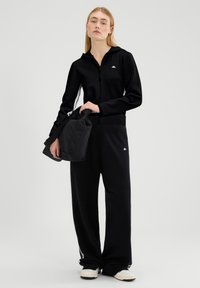 Czarna bluza z kapturem na zamek błyskawiczny z prążkowanymi mankietami, zestawiona z luźnymi czarnymi spodniami z białym logo. Model trzyma czarną torbę. Prosty design.