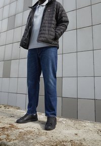 Man som bär en svart jacka, vit polo, blå jeans och svarta sneakers står på grus framför en grå kaklad vägg.
