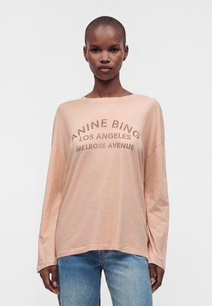Osoba stojí, oblečená v voľnej svetloružovej košeli s dlhými rukávmi s textom "ANINE BING LOS ANGELES MELROSE AVENUE" a v modrých džínsoch.
