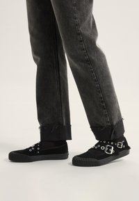 Zapatillas de lona negras con acentos de hebilla plateada, parte superior texturizada y suela de goma. Combinadas con jeans de mezclilla negros desgastados y con dobladillo.