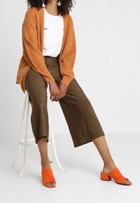 Cardigan naranja con relieves, camiseta gráfica blanca, culottes de rayas marrones, y sandalias bloque de ante naranja, sentada en un taburete blanco.