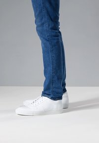 Zapatillas deportivas de cuero blanco con textura suave, diseño de perfil bajo y costuras en contraste, usadas con vaqueros azul ajustados.