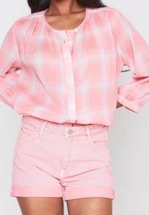 Kobieta ubrana w różowo-białą kraciastą koszulę na guziki z rękawami długości 3/4 oraz jasnoróżowe, podwinięte szorty z denimu.