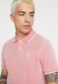 Rød polo skjorte med en tekstureret stof, hvide accenter på kraven og et rundt logo på brystet. Standard pasform og korte ærmer.