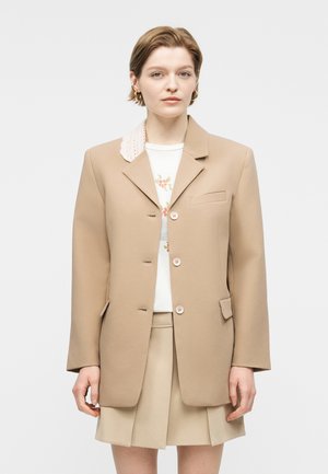 Γυναίκα με κοντά μαλλιά που φοράει μπεζ σακάκι πάνω από λευκό φλοράλ τοπ και μπεζ πλισέ φούστα, στέκεται μπροστά σε απλό φόντο.