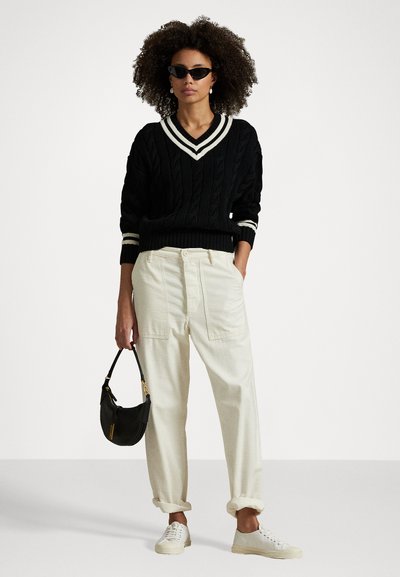Pull en maille torsadée noir avec des rayures blanches au col, pantalon couleur crème, sac à main noir et baskets blanches. Le modèle porte des lunettes de soleil noires.