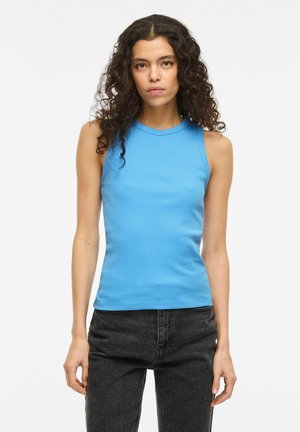 Kobieta o kręconych ciemnych włosach, ubrana w bezrękawnikowy niebieski prążkowany top i czarne dżinsy, stojąca na jednolitym białym tle.