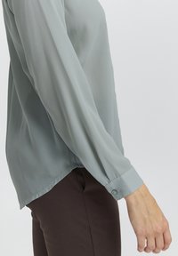 Camicia leggera blu chiaro, trasparente, con maniche lunghe e polsini abbottonati. Indossata con pantaloni fitted marrone scuro, con orlo curvo.