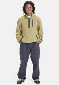 Fleece pullover in licht olijfgroen met een grijze paneel, knopen en zijzakken. Draagt loszittende marineblauwe broek en donkere sneakers.