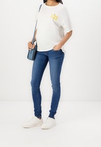 Vitt T-shirt med "MOM PWR" i gult, tillsammans med figurnära blå jeans och vita sneakers. En liten blå crossbody-väska kompletterar looken.