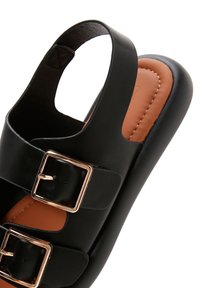Black sandal featuring two wide straps with gold buckles, an open back design, and a brown cushioned insole. Smooth leather-like texture.