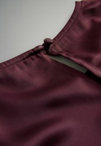 Bordeaux stof med en glat tekstur; har en rund halsudskæring og en dekorativ knaplukning bagpå.