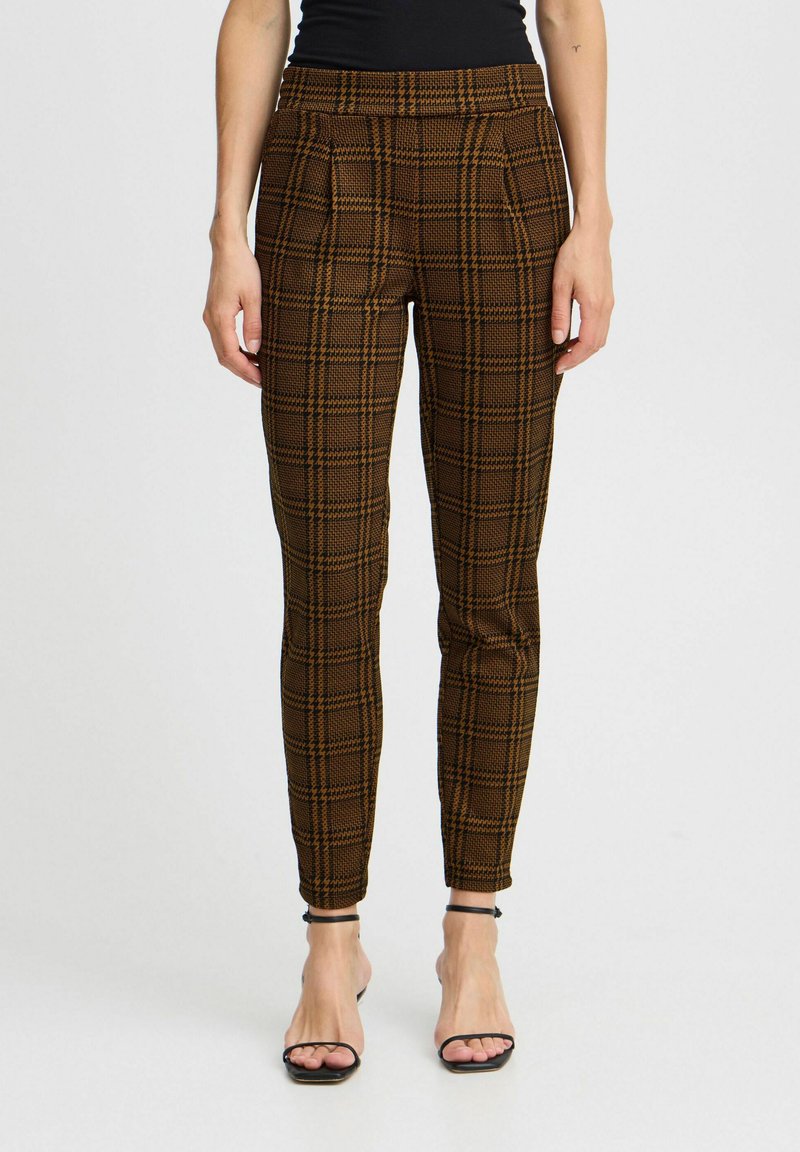 Braune karierte Hose im Slim-Fit-Schnitt mit hoher Taille, Falten auf der Vorderseite und einem strukturierten Stoff. Sichtbare schwarze Riemchensandalen.