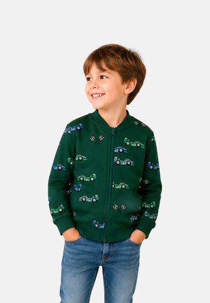 Junger lächelnder Junge, der eine grüne Jacke mit Reißverschluss und blauen und grünen Rennwagen-Motiven sowie blaue Jeans trägt, Hände in den Taschen, schaut zur Seite.